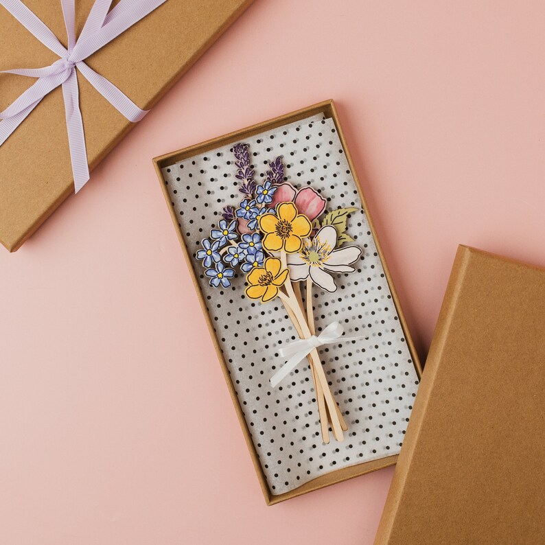 Op de afbeelding: Een boeket van papieren bloemen in tinten geel, blauw en roze, met een wit lint om de stelen gebonden. De bloemen zijn gerangschikt in een doos met stippen.