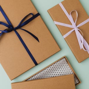May include: Three brown gift boxes with different colored ribbons. The top box has a blue ribbon tied in a bow. The middle box has a purple ribbon tied in a bow. The bottom box is open and has a white and black polka dot lining.