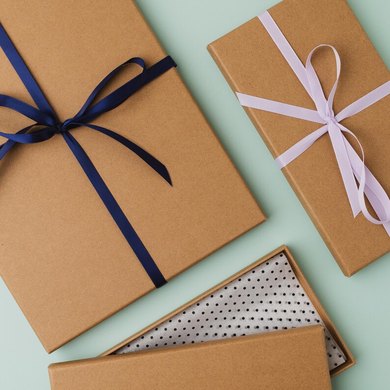 May include: Three brown gift boxes with different colored ribbons. The top box has a blue ribbon tied in a bow. The middle box has a purple ribbon tied in a bow. The bottom box is open and has a white and black polka dot lining.