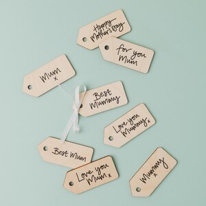 May include: Eight wooden gift tags with handwritten messages for Mother's Day. The tags are light brown and have black ink. The messages include "Mum", "Best Mummy", "Love you Mummy", "Happy Mother's Day", "For you Mum", and "Best Mum".