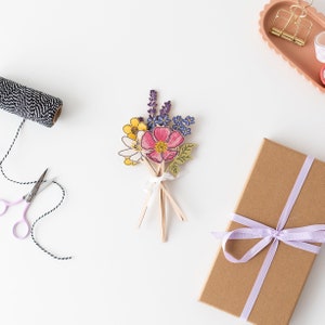 Op de afbeelding: Een boeket van papieren bloemen in tinten roze, geel, paars en groen. De bloemen zijn in een bosje gerangschikt en met een wit lint bij elkaar gebonden. Het boeket ligt op een wit oppervlak.