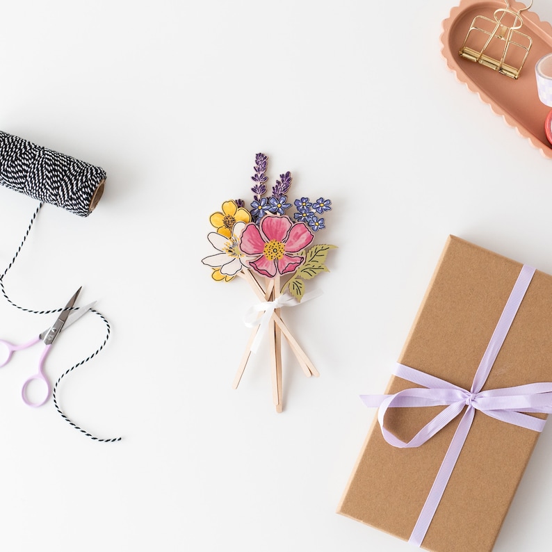 Op de afbeelding: Een boeket van papieren bloemen in tinten roze, geel, paars en groen. De bloemen zijn in een bosje gerangschikt en met een wit lint bij elkaar gebonden. Het boeket ligt op een wit oppervlak.