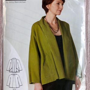 May include: A sewing pattern envelope for an Indygo Essentials Swing Jacket by Amy Barickman, featuring a model wearing an olive green open-front jacket with wide sleeves and front pockets, alongside three black and white line art sketches.