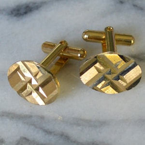 May include: A pair of gold cufflinks with a geometric pattern. The cufflinks are oval shaped and have a textured surface.