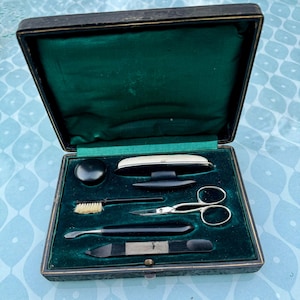 May include: An open, antique black leather case with a green velvet lining. The case contains grooming tools, including a brush, scissors, and nail tools. The case is rectangular with a hinged lid.