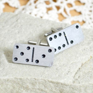 May include: A pair of silver domino cufflinks. Each cufflink features a silver domino with black dots. The domino on the left cufflink has three dots on one side and one dot on the other side. The domino on the right cufflink has four dots on one side and one dot on the other side.