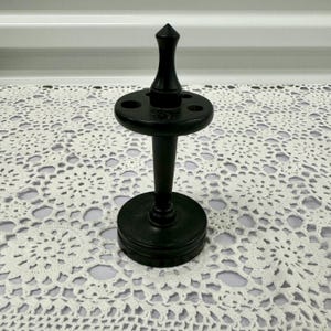 Op de afbeelding: Een zwarte houten tandenborstelhouder. Het heeft een ronde basis, een centrale paal en een cirkelvormige bovenkant met vier gaten. De bovenkant heeft een puntige bekroning. De houder staat op een wit kanten kleedje.