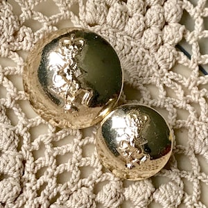 May include: Two gold-colored buttons with a raised design of a crown and a figure holding a sword. The buttons are on a white crocheted surface.