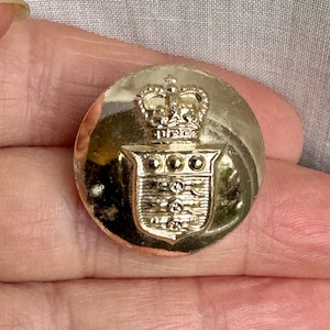 Puede incluir: Botón dorado con un diseño de corona y escudo. La corona tiene tres puntas y el escudo tiene tres bandas horizontales.