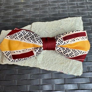 May include: A bow tie with a white, black, yellow, and burgundy striped pattern. The bow tie is tied and ready to wear.
