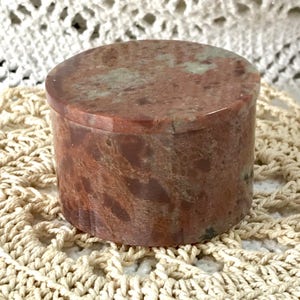 Puede incluir: Una caja de piedra redonda, roja, verde y marrón con tapa. La caja está hecha de piedra natural y tiene un acabado liso y pulido.