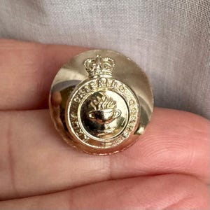 May include: A gold-colored button with a crown and the words "Royal Catering Corps" around a stylized image of a cup with a flame above it.