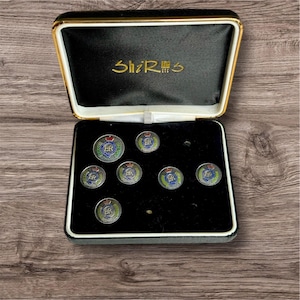 Puede incluir: Una caja de terciopelo negro con ribete dorado que contiene nueve gemelos dorados con un diseño de esmalte azul, rojo y verde. Los gemelos presentan las letras "ER" en el centro. La caja tiene el texto "ShiR  ETC" en la tapa.