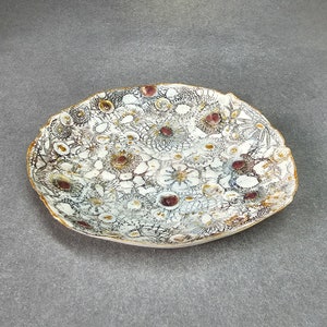 Könnte beinhalten: Eine weiße Keramik-Servierplatte mit einem gewellten Rand. Die Platte ist mit einem Muster aus kleinen weißen Blüten und braunen und violetten Punkten verziert.