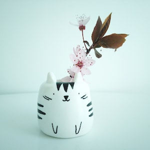 Può includere: Un vaso di ceramica bianco a forma di gatto con strisce nere e un viso carino, che tiene un ramo di fiori di ciliegio rosa.