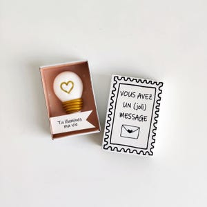 Puede incluir: Un objeto blanco en forma de bombilla con un corazón y una base dorados, dentro de una caja rosa. La caja tiene una pancarta blanca con el texto "Tu illumines ma vie". Al lado hay una tarjeta blanca con borde negro y el texto "VOUS AVEZ UN (joli) MESSAGE".