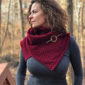 May include: A woman wearing a burgundy knitted cowl neck scarf with a gold metal pin. The scarf is textured and has a triangular shape.