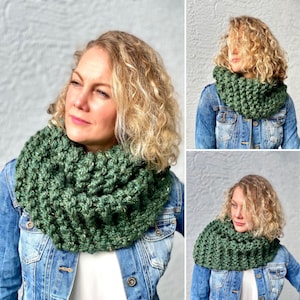 May include: A green crocheted infinity scarf worn by a person with curly blonde hair. The scarf is thick and textured, and the person is wearing a denim jacket.