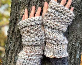 May include: A pair of knitted fingerless gloves in a beige and brown color with a speckled pattern. The gloves are made with a chunky yarn and have a ribbed cuff.