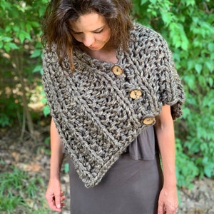Puede incluir: Un poncho de punto marrón con botones de madera. El poncho se lleva sobre un vestido gris.