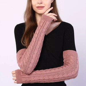 May include: A pair of pink knitted arm warmers with a cable knit pattern. The arm warmers are worn over a black sweater with a V-neck.