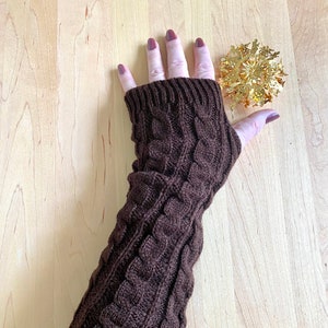 May include: Brown knitted arm warmers with a cable knit pattern. The arm warmers are on a light wood surface.