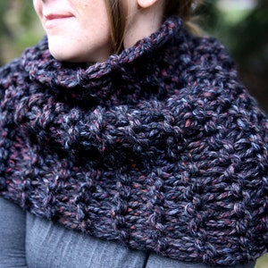 May include: A close-up of a dark gray, black, and pink knitted cowl neck scarf. The scarf is made of a chunky yarn and has a textured pattern.