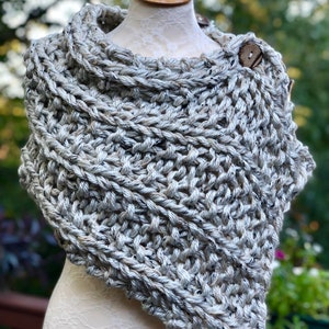 May include: A grey knitted shawl with a chunky texture and a wooden button. The shawl is draped over a white mannequin.