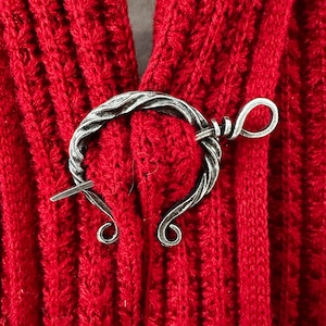 Puede incluir: Un broche de chal de metal plateado con un diseño retorcido y un bucle en el extremo. El broche está sobre una tela de punto roja.