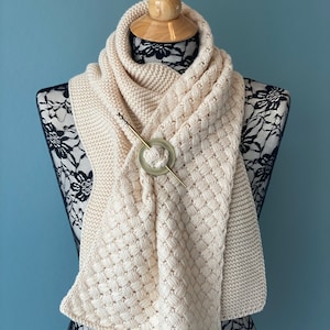 Peut inclure: Une écharpe en tricot de couleur crème avec un motif tissé et une épingle dorée avec un élément décoratif rond.