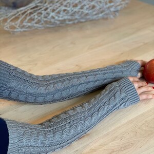 May include: A pair of gray knitted arm warmers with a cable knit pattern. The arm warmers are shown on a wooden table.