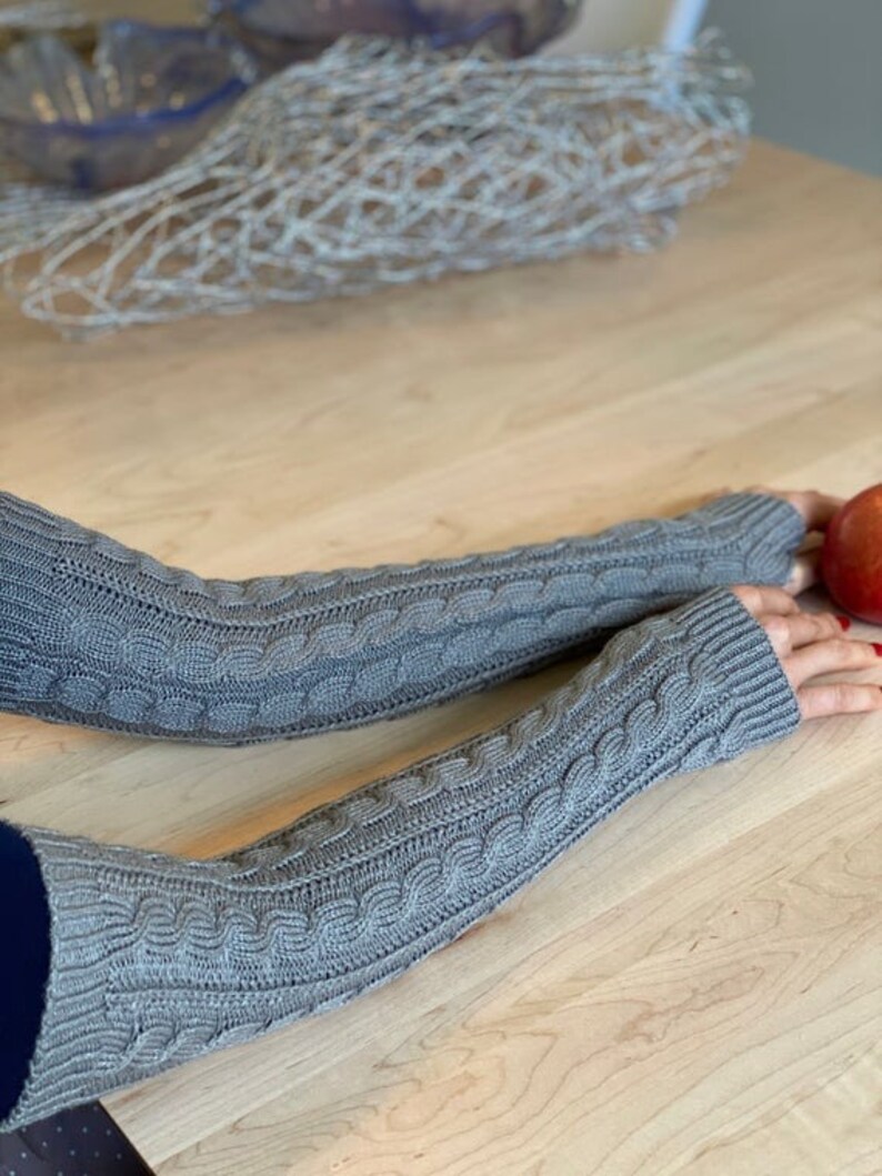 May include: A pair of gray knitted arm warmers with a cable knit pattern. The arm warmers are shown on a wooden table.