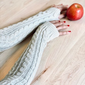 May include: A pair of beige knitted arm warmers with a cable knit pattern. The arm warmers are on a wooden surface with a red apple.