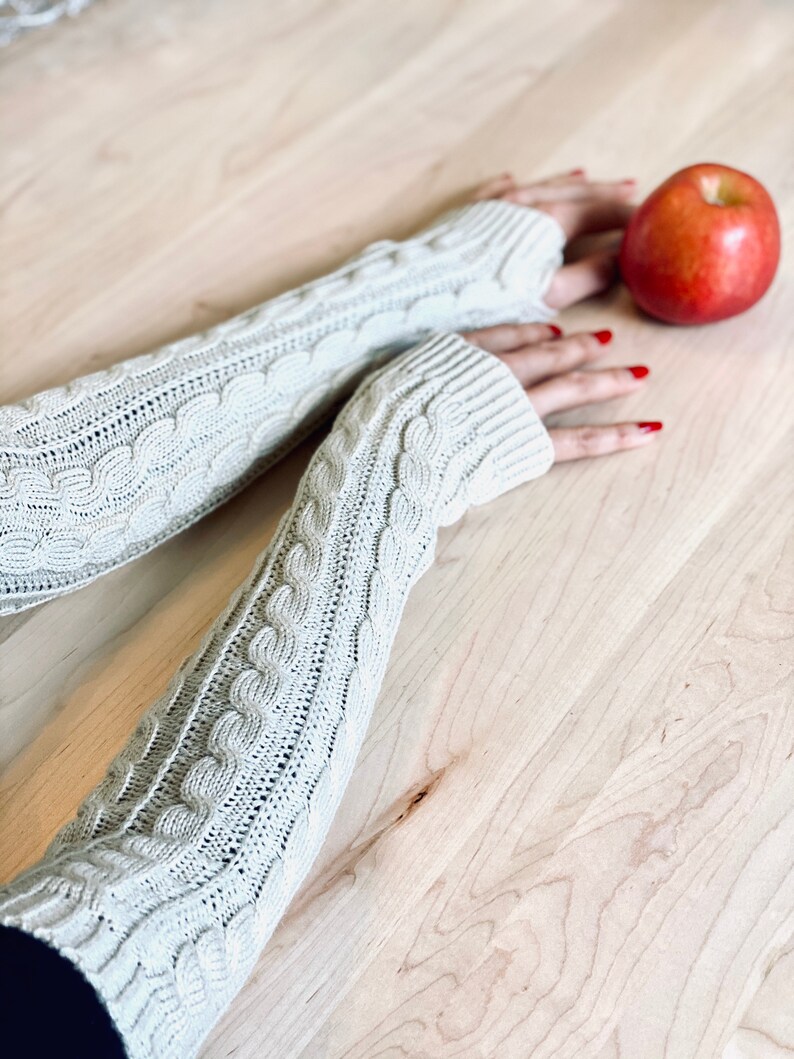 May include: A pair of beige knitted arm warmers with a cable knit pattern. The arm warmers are on a wooden surface with a red apple.