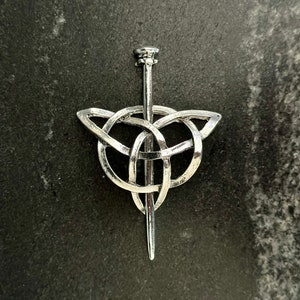 Puede incluir: Broche de nudo celta plateado con un alfiler central. El broche presenta un intrincado diseño entrelazado, con una forma triangular en la parte superior y un alfiler puntiagudo que se extiende hacia abajo. El metal tiene un acabado pulido, que refleja la luz.