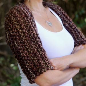 May include: A brown and cream knitted shrug with a loose fit. The shrug is made with a chunky yarn and has a simple, elegant design.