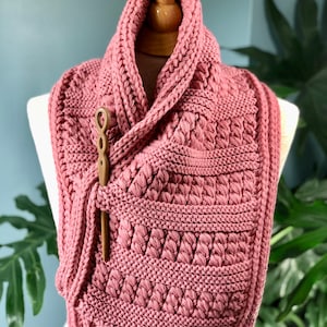Puede incluir: Una bufanda de cuello de tortuga de punto rosa con una aguja de tejer de madera metida en el pliegue.