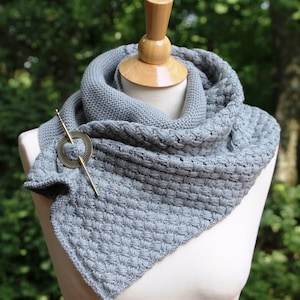 May include: A gray knitted infinity scarf with a decorative pin. The pin is made of metal and has a circular design.
