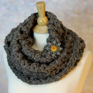 May include: A gray crocheted infinity scarf with a decorative flower brooch featuring a yellow button.
