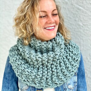May include: A woman wearing a light blue knitted cowl neck scarf. The scarf is made with a chunky yarn and has a textured stitch pattern.