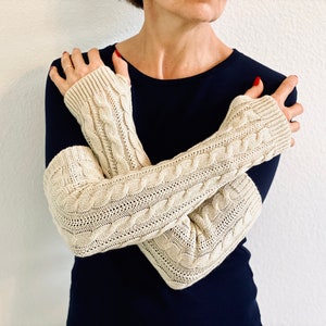 May include: A pair of cream-colored knitted arm warmers with a cable knit pattern. The arm warmers are worn by a person with a dark blue shirt.