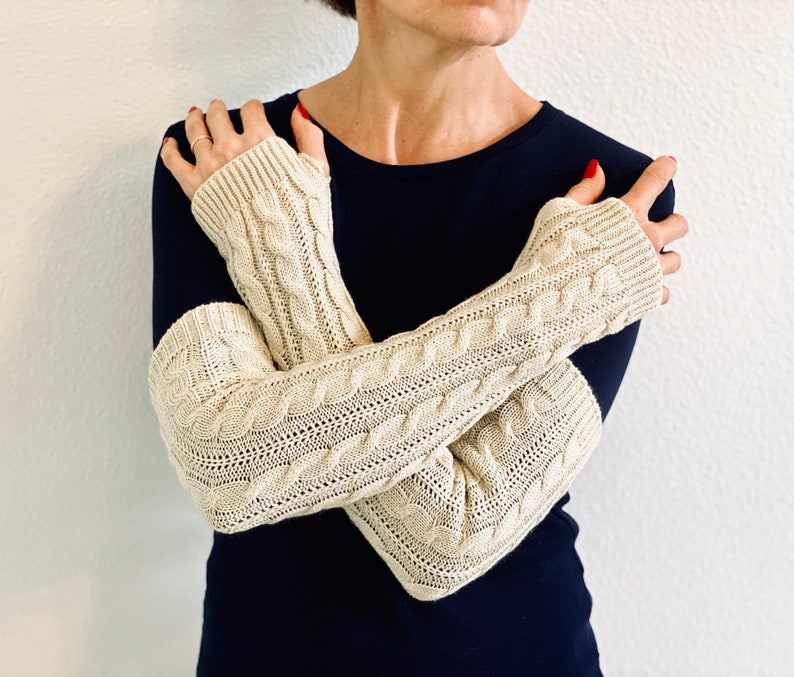 May include: A pair of cream-colored knitted arm warmers with a cable knit pattern. The arm warmers are worn by a person with a dark blue shirt.