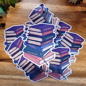 以下が含まれることがあります： 「ストーン・チェンバー」「アズカバン」「ゴブレット」「フェニックス」「ハーフブラッド」「ハロウズ」というタイトルの書かれた本が積み重ねられたステッカーが複数あります。ステッカーは木製の表面に貼られています。