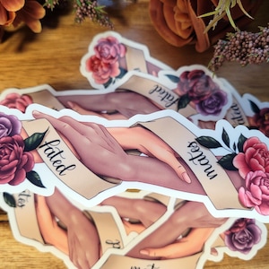 May include: Two hands intertwined with the words "fated" and "lovers" on a banner with pink and purple flowers.