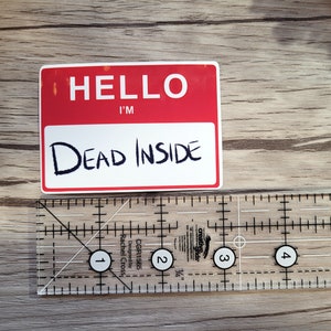 May include: A white sticker with red lettering that reads "Hello I'm Dead Inside". The sticker is on a wooden surface.