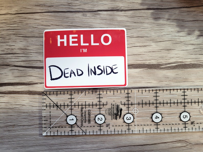 May include: A white sticker with red lettering that reads "Hello I'm Dead Inside". The sticker is on a wooden surface.