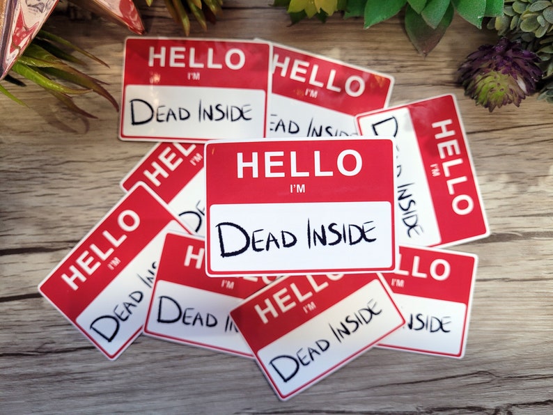May include: A stack of red and white name tag stickers with the text "Hello I'm Dead Inside" printed on them. The stickers are on a wooden surface.