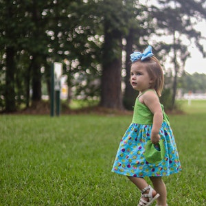 May include: A young girl wearing a green and blue dress with a light blue bow in her hair. She is standing in a grassy field and looking to the right.