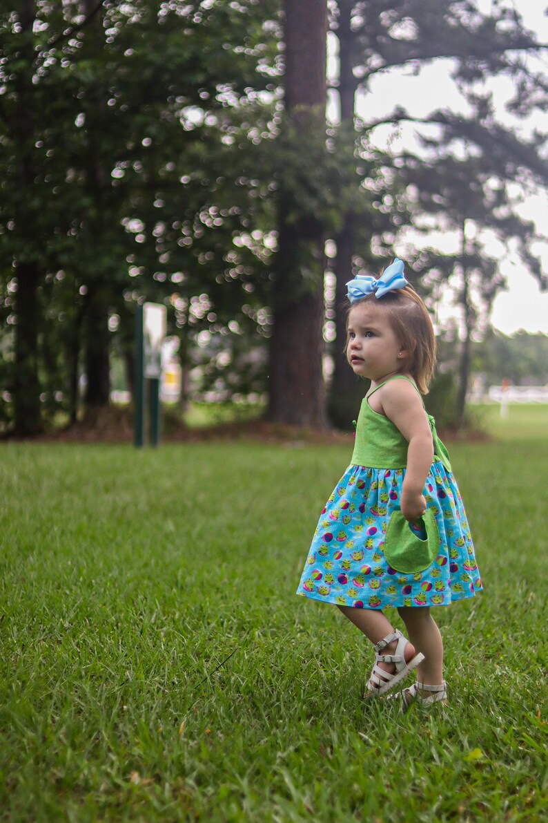 May include: A young girl wearing a green and blue dress with a light blue bow in her hair. She is standing in a grassy field and looking to the right.