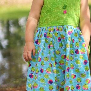 May include: A young girl wearing a green and blue sundress with a frog and beach ball pattern. The dress has a green bodice with a white polka dot pattern and a blue skirt with a frog and beach ball pattern.